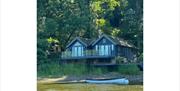 Lakeside wooden cabin with glass balcony overlooking Rudyard Lake, with a small rowboat on the water in the foreground.