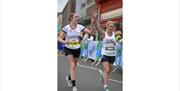 Runners crossing a city centre finish line, raising arms in celebration during a road race event.