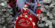 Image 1: Festive scene with two Christmas elves sitting on a red North Pole Mail barrel surrounded by snow-covered trees.
Image 2: Father Christmas se