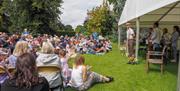 An outdoor theatre performance, with the cast in a marquee and a large crowd of people sitting in garden chairs or on the lawn