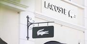 A hanging black sign featuring the Lacoste crocodile symbol mounted, with the Lacoste store logo displayed on the building behind it.