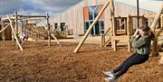 A child using a rope swing in an outdoor adventure playground with wooden climbing structures and bark flooring. A large modern wooden building sits i