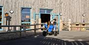 An adult and two children riding a large outdoor pedal cart along a paved path outside a wooden building with blue-framed doors and windows. Morning s