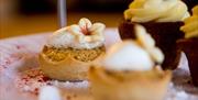 A close-up of delicate mini desserts, including a tart topped with cream and a small flower decoration, arranged on a plate with crumbs and powdered g