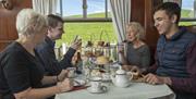 A family on a classic steam train tuck into an afternoon cream tea. With scones and cakes on a tower, and teapots and bottled beers on the table.
