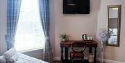 Daylight streams into the bedroom through the large window. You can see a long mirror on the wall, as well as a corner of the bed, and the TV, desk an